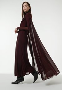 Bordeauxfarbendes Kleid mit durchsichtigen Umhangärmeln und hohem Kragen, kombiniert mit schwarzen spitzen Knöchelstiefeln, die eine fließende Silhouette betonen.