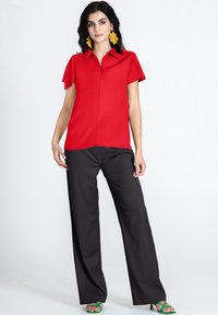 Blusa rossa a maniche corte con colletto e zip frontale abbinata a pantaloni neri a gamba larga. La modella indossa sandali verde con cinturini e grandi orecchini gialli.