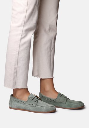 Grüne Mokassins aus Wildleder, getragen mit kurz geschnittenen, cremefarbenen Hosen vor einem schlichten hellen Hintergrund.