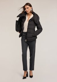 Giacca a vento nera con colletto alto, indossata sopra una blusa chiara. Abbinata a jeans scuri e tacchi a punta neri, con una grande borsa a tracolla.