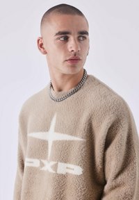 Jersey de color beige, suave y difuso, con una estrella blanca y el logo "PXP". Combinado con un collar de cadena plateada, mostrando una apariencia texturizada.