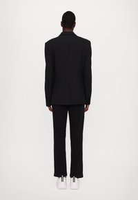 Blazer nero su misura con spalle definite, abbinato a pantaloni neri coordinati e sneakers bianche con strisce nere.