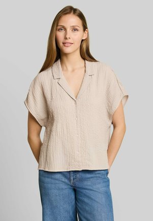 Jonge vrouw met lang bruin haar, die een beige kortemouwen gestructureerd overhemd met knopen en een blauwe spijkerbroek draagt, staat met haar handen achter haar rug.
