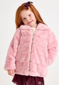 Giacca di pelliccia finta rosa con colletto, chiusura con zip e texture trapuntata. Indossata sopra una gonna bordeaux, con maniche lunghe.