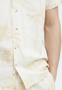 Short-sleeve linen shirt in off-white with a subtle palm tree pattern in light beige, featuring a chest pocket and button closure.