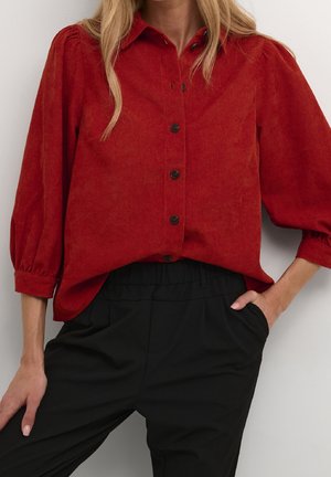 Femme portant une chemise rouge boutonnée à manches longues bouffantes et un pantalon noir taille haute, une main dans la poche.