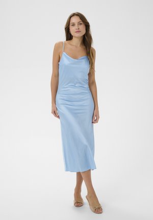 Nő világoskék szatén, vékony pántos csúszkában, bézs nyitott orrú magas sarkúban, egyszínű fehér háttér előtt áll.