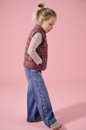 Lite barn med blondt hår i hestehale iført vinrød quiltet vest, stripete langermet skjorte, vide bukser med hjerte-design, og beige sko.