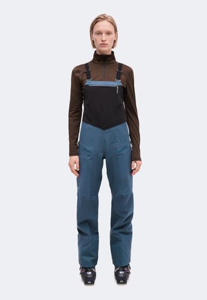 Le modèle se tient face à l’objectif, vêtu d’un haut marron à col zippé, d’une bavette noire, d’un pantalon de ski bleu et de chaussures de ski, sur un fond clair uni.