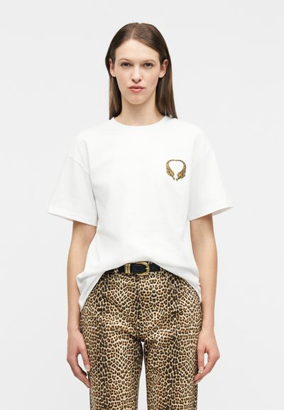 Femme portant un t-shirt blanc oversize avec un petit motif de tigre et un pantalon imprimé léopard maintenu par une ceinture noire avec une boucle en laiton.