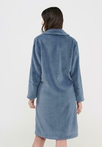 Mujer con una bata azul texturizada de manga larga y hasta la rodilla, de pie con su espalda a la cámara contra un fondo liso.