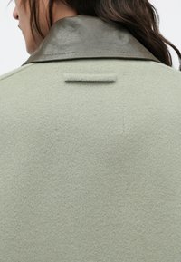 Zeleni plašč iz volnenega mešanega materiala z rjavim usnjenim ovratnikom, z zanko na hrbtni strani in mehko teksturo. Čiste linije in minimalističen dizajn.