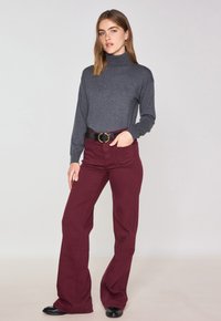 Maglione a collo alto grigio scuro abbinato a pantaloni a campana bordeaux, cintura nera con fibbia circolare dorata e scarpe nere su uno sfondo neutro.
