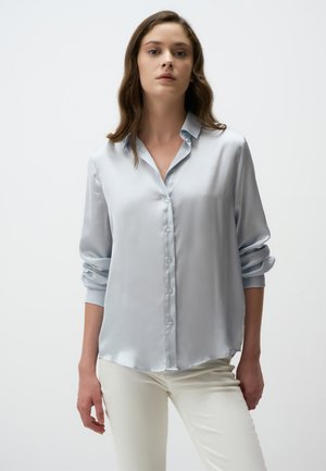 Light blue satin blouse with a button-down front, long sleeves rolled up, and a curved hem. Smooth texture and subtle sheen.