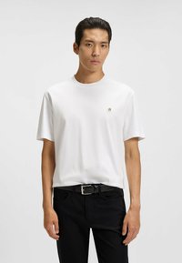 T-shirt bianca a maniche corte realizzata in tessuto morbido, con un piccolo logo sul petto, abbinata a jeans neri e una cintura nera.