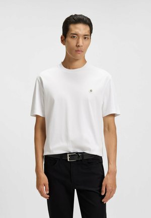 Weißes kurzärmeliges T-Shirt aus glattem Stoff, mit einem kleinen Logo auf der Brust, kombiniert mit schwarzen Jeans und einem schwarzen Gürtel.