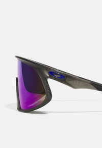 Ochelari de soare cu ramă neagră și lentile cu nuanțe de violet. Prezintă un design elegant și accente texturate aproape de tâmple pentru un plus de aderență.