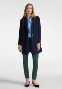 Abrigo de lana azul marino con un solo botón, camisa de cuello azul claro, pantalones verdes y zapatos de punta marrón oscuro. Textura suave en todo.