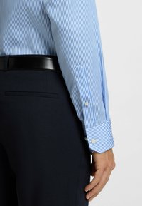 Camicia a righe blu e bianche con polsini abbottonati, abbinata a una cintura nera e pantaloni blu scuro. Tessuto liscio con una vestibilità su misura.