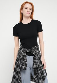 Sort kortærmet t-shirt lavet af ribstrikket stof, kombineret med en sort og gråternet skjorte bundet om livet over lysegrå bukser.