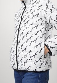 Vit puffjacka med svart handskriven "Hugo"-mönster, med dragkedja fram, mjuk textur och svarta detaljer runt ärmslut och nederkant.