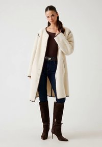 Langer, cremefarbener Mantel aus weichem, strukturiertem Stoff mit dunklem Besatz. Trägt man über einem braunen Oberteil, kombiniert mit eng anliegenden dunklen Jeans und braunen Stiefeln.