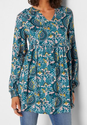 Femme portant un chemisier à manches longues fleuri couleur sarcelle avec des motifs roses, jaunes et blancs, et un jean bleu, debout devant un fond blanc.