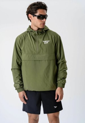 Man wearing black sports sunglasses, an olive green hooded jacket with "Toujours Actif" text, and black shorts with matching branding stands against a plain background.