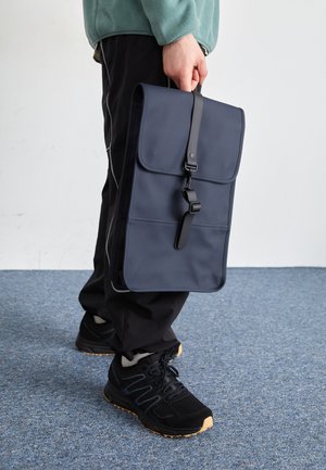 Navy blue foldable bag with a smooth surface, secured by a black strap. A person holds it while wearing black athletic shoes and dark pants.