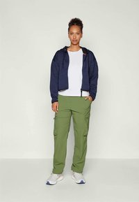 Felpa con zip color navy in tessuto testurizzato, t-shirt bianca, pantaloni cargo oliva con tasche e sneakers bianche con dettagli blu.