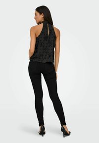 Femme aux longs cheveux noirs portant un haut noir sans manches à motif floral, un jean skinny noir et des chaussures à talons hauts noires, vue de dos.