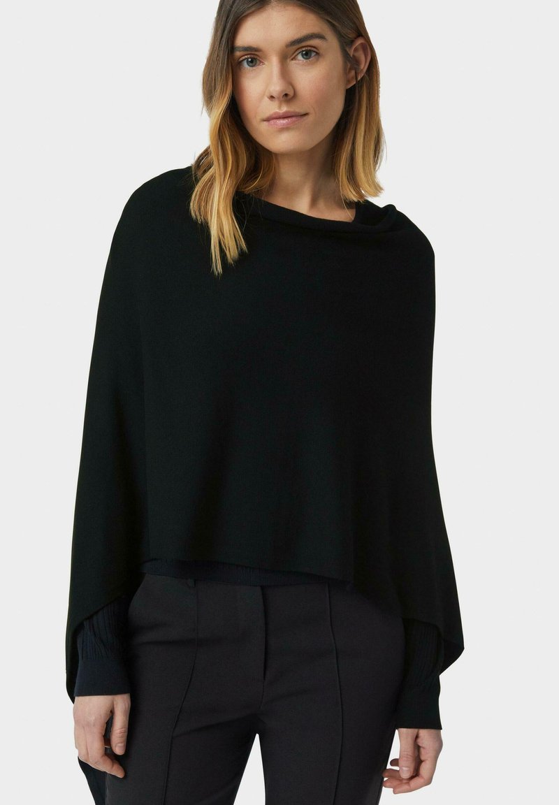 Donna con capelli biondi alla spalla, che indossa un maglione nero a poncho e pantaloni neri, in piedi contro uno sfondo chiaro e semplice.