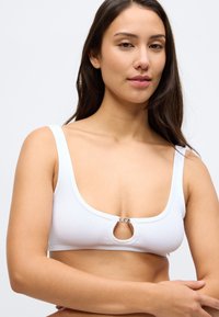 Λευκό ριμπ crop top με τετράγωνη λαιμόκοψη και κυκλικό μεταλλικό διακοσμητικό στη μέση. Ρυθμιζόμενες τιράντες με εφαρμοστή γραμμή.