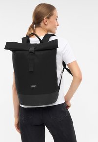 Sac à dos noir à rabat, fabriqué en tissu durable, avec des sangles ajustables et une forme rectangulaire. Présente un logo en façade.