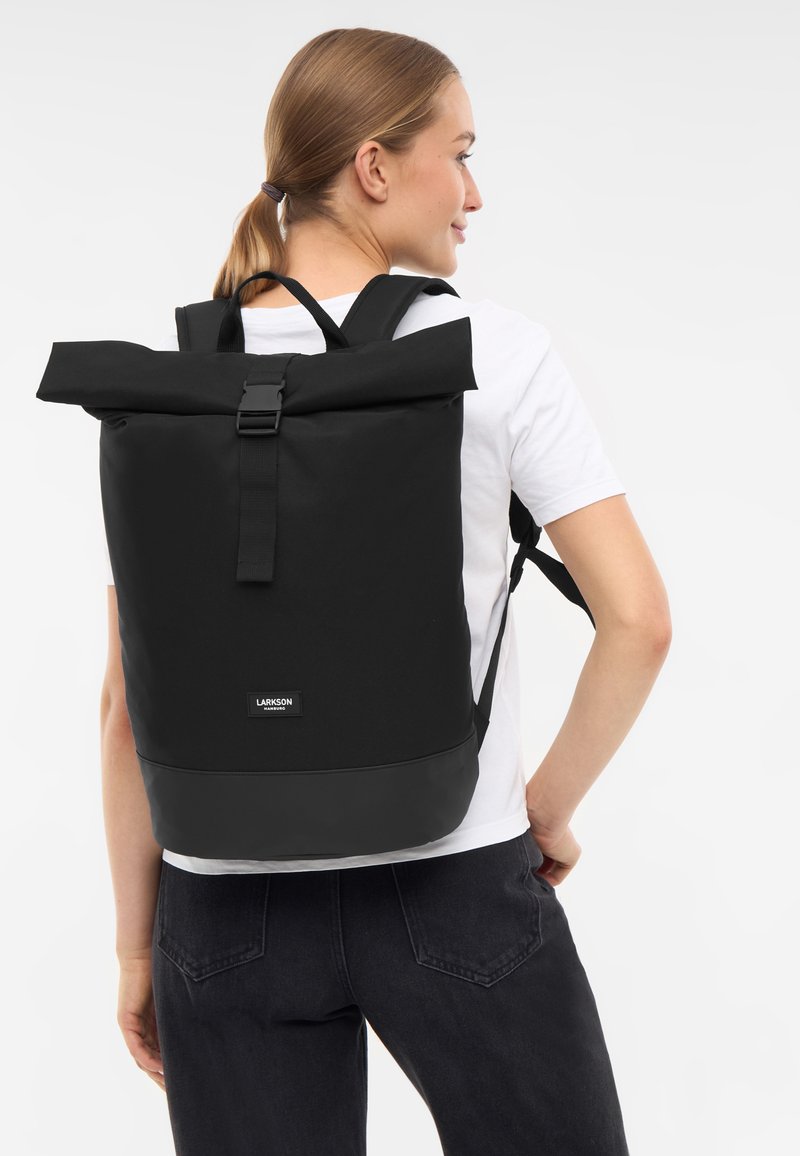 Sac à dos noir à rabat, fabriqué en tissu durable, avec des sangles ajustables et une forme rectangulaire. Présente un logo en façade.