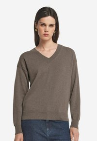 V-Ausschnitt, langärmliger Pullover in einem gedämpften Braunton, aus weichem Strickstoff mit gerippten Bündchen und Saum. Kombiniert mit dunkler Denim.
