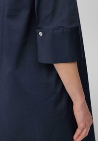 Marineblaue Hemd in lockerer Passform, mit einem umgeschlagenen Ärmel, der mit einem grauen Knopf gesichert ist. Glattes Baumwollmaterial, minimalistisches Design.