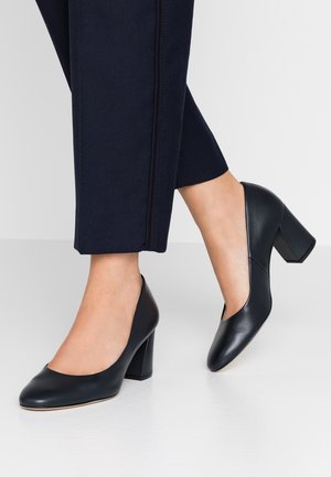 Zwarte leren pumps met een ronde neus en blokhak, gedragen bij marineblauwe broeken, met een gladde textuur en eenvoudig ontwerp.