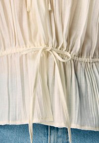 Blouse à rayures blanches avec une taille cintrée, dotée d'un lien en ruban. Le tissu léger a une texture douce et un design fluide.