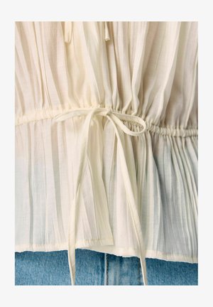 Bluză albă cu dungi, cu talie strânsă, având un șnur legat. Materialul ușor are o textură moale și un design fluid.