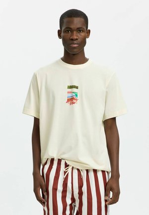 Jeune homme portant un T-shirt crème avec un texte graphique coloré « Leisurely Vibe » et un short rayé vertical rouge et blanc, debout sur fond blanc.