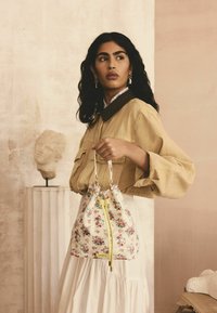 Blomsterbucket taske i creme med lyserøde roser, guldhardware og en limegrøn snor. Båret af en model i en beige bluse.