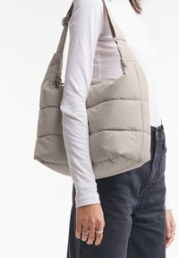 Borsa a spalla beige imbottita con una texture morbida e trapuntata, con tracolla regolabile, tenuta da una persona che indossa una camicia a maniche lunghe bianca.