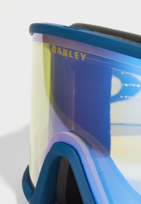 Niebieskie gogle narciarskie z gradientowymi purpurowymi soczewkami i złotym logo "Oakley". Posiadają wyściółkę z pianki i zakrzywiony kształt ramy dla wygody.
