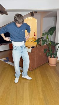 Ανοιχτού μπλε φθαρμένα τζιν με άνετη εφαρμογή, φορεμένα με μια σκουρόχρωμη υφασμάτινη μπλούζα. Εσωτερικός χώρος με μια γλάστρα και καφέ καναπέ.