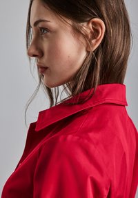 Červená bunda s límcem, hladkým povrchem, s viditelným švem a elegantním designem, kterou nosí modelka v bočním profilu na neutrálním pozadí.