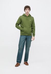 Πράσινο βαμβακερό φούτερ με κουκούλα και ρυθμιζόμενα κορδόνια, τσέπη μπροστά και ριμπ μανσέτες, συνδυασμένο με μπλε τζιν και καφέ καραβόπανα παπούτσια. Απλό φόντο.