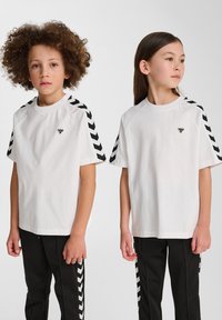 T-shirt blanche avec des accents en chevrons noirs sur les manches, associée à un pantalon noir avec des rayures latérales similaires. Design minimaliste.
