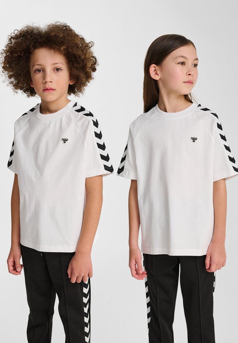 T-shirt blanche avec des accents en chevrons noirs sur les manches, associée à un pantalon noir avec des rayures latérales similaires. Design minimaliste.