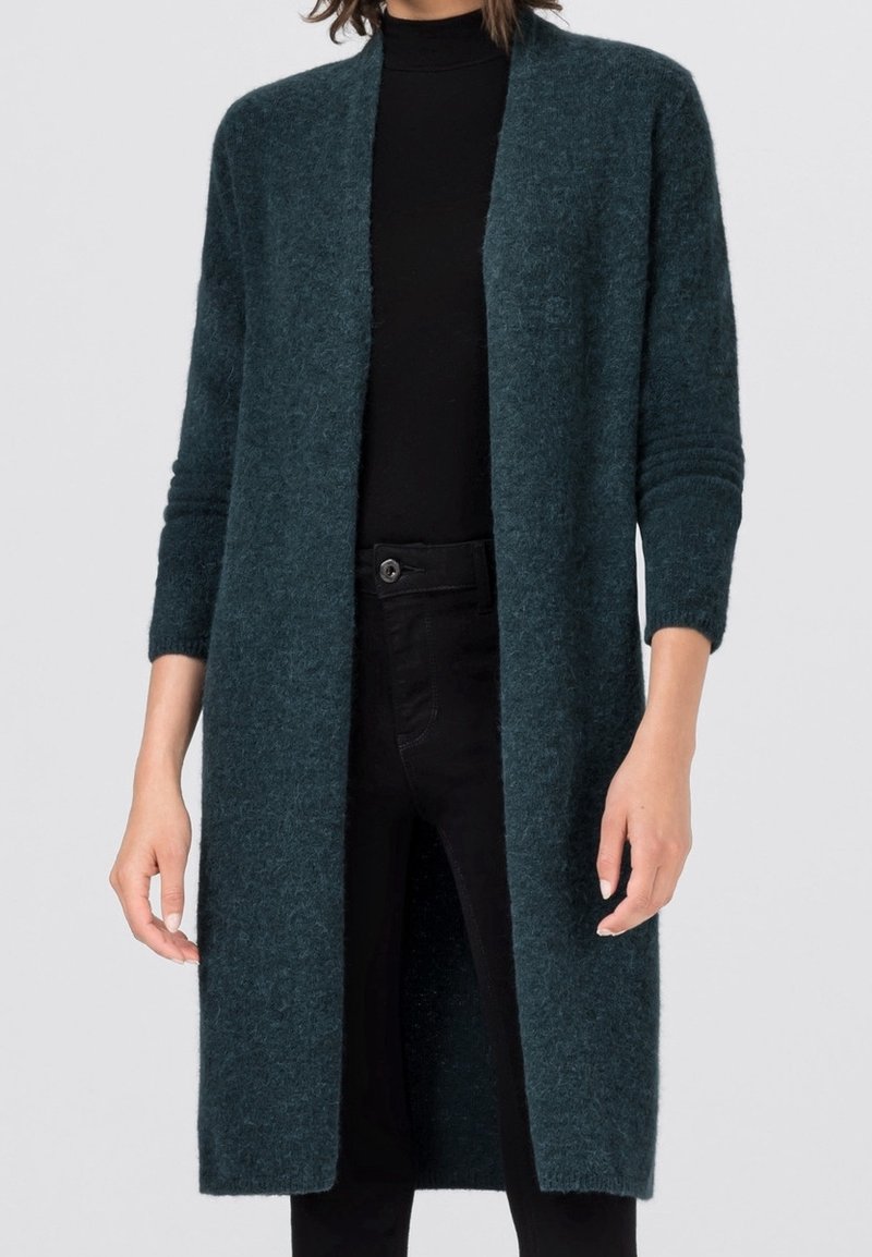 Femme portant un cardigan long à ouverture frontale de couleur bleu sarcelle foncé, sur un col roulé noir et un pantalon noir, debout devant un fond uni.